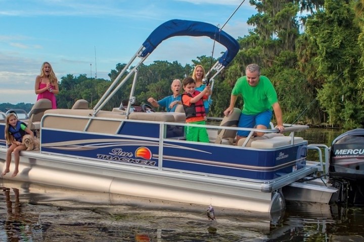 Family on a pontoon boat fishing and relaxing on a lake with trees in the background.